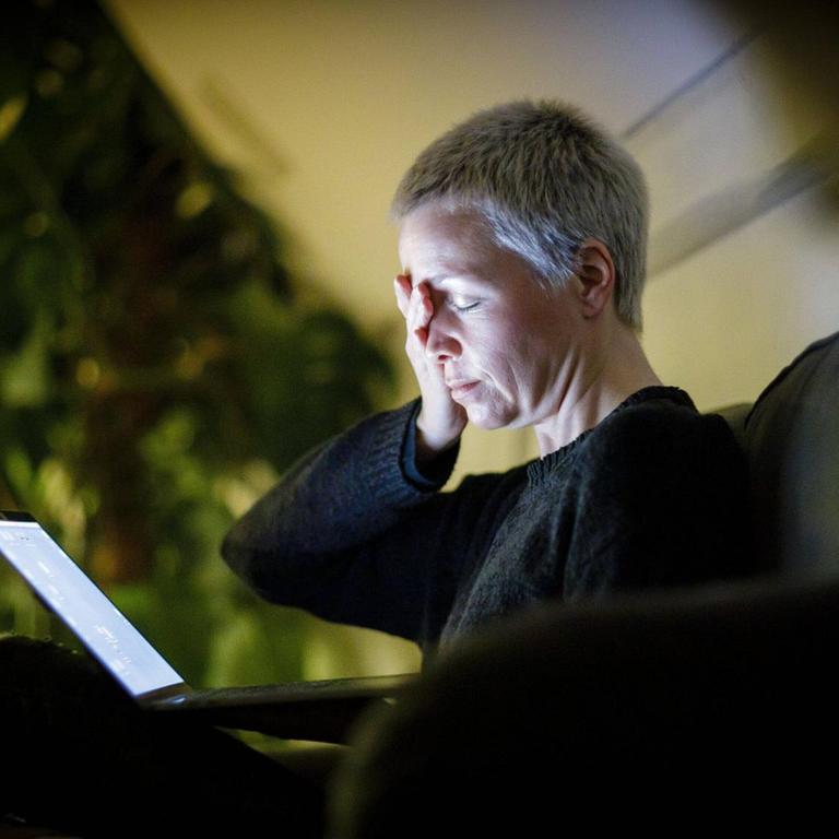 Eine Frau sitzt mit geschlossenen Augen auf dem Sofa vor ihrem Laptop und hat ihre Hand auf die Stirn gelegt. Symbolbild.