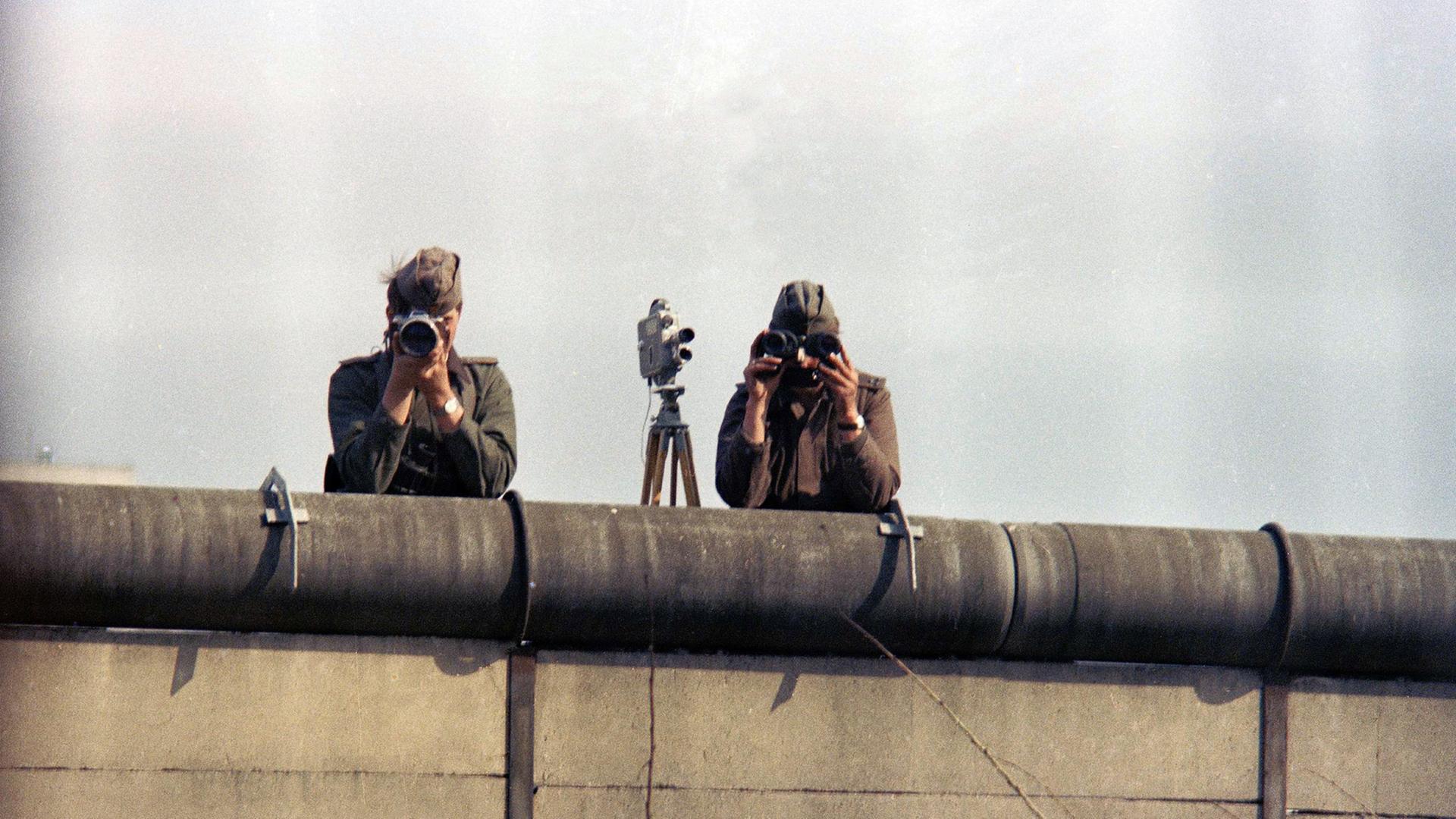 1976: Zwei Ostberliner Grenzposten fotografieren von der Mauer aus Menschen in Westberlin.