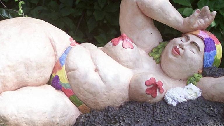 Skulptur einer Badenixe von der Künstlerin Leda Astorga, die eine füllige, liegende Frau beim Sonnenbaden zeigt, wobei über ihren Brustwarzen große, rote Blumen gelegt sind und sie eine bunte Badekappe trägt.