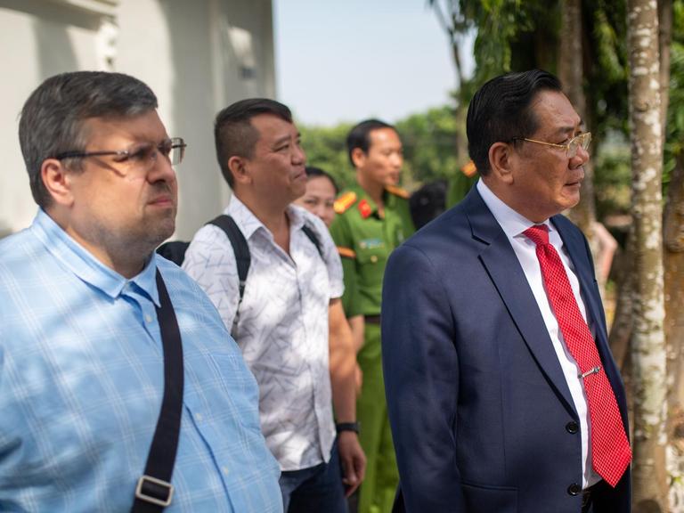 Der Generalleutnant Ho Thanh Dinh in blauem Anzug und roter Krawatte, hinter ihm laufen zwei Journalisten.