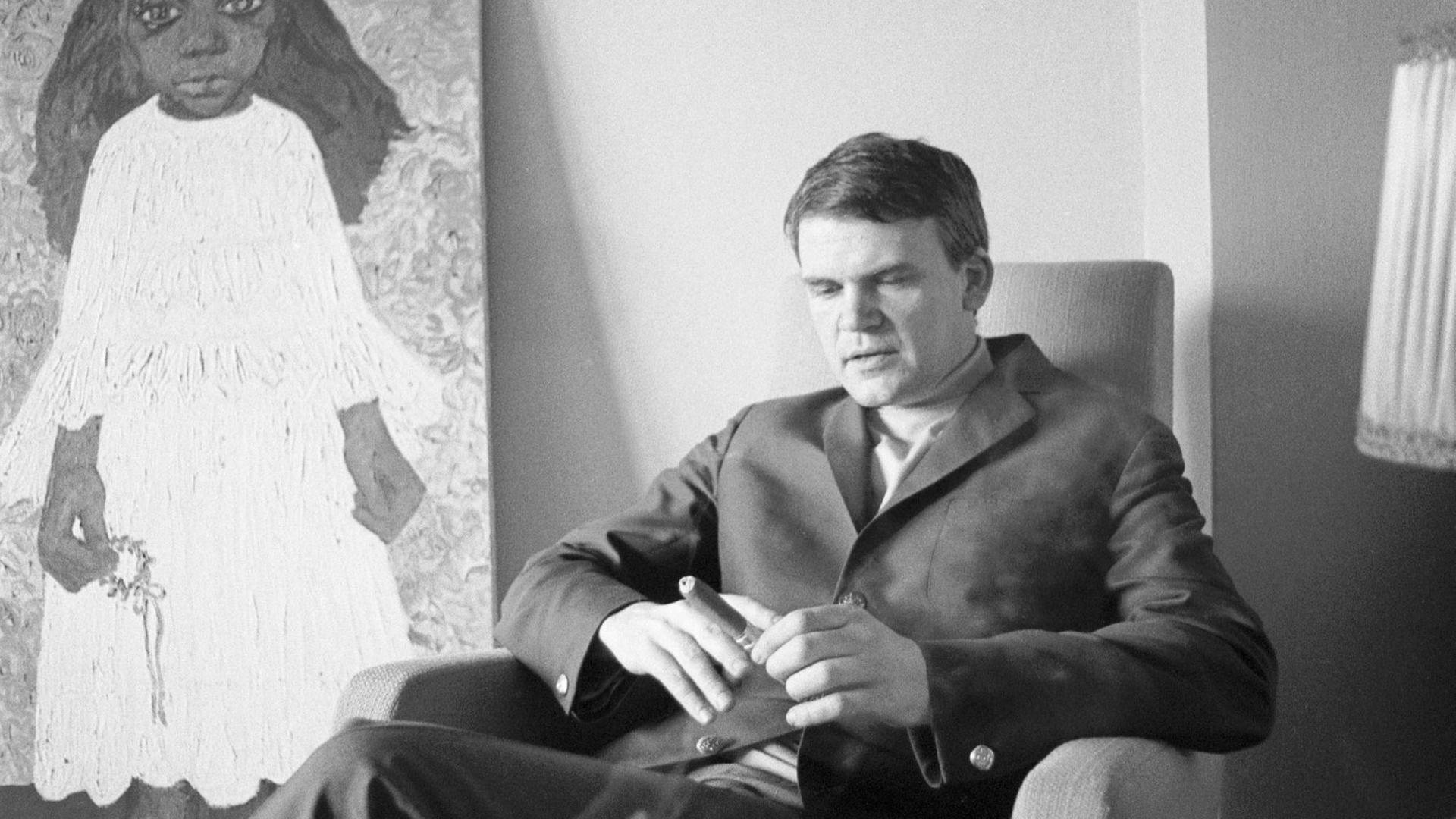 Milan Kundera sitzt mit einer Zigarre in der Hand auf einem Sessel. Rechts von ihm steht eine Lampe, links von ihm steht das Gemälde eines Mädchens. Die Aufnahme ist schwarz weiß.