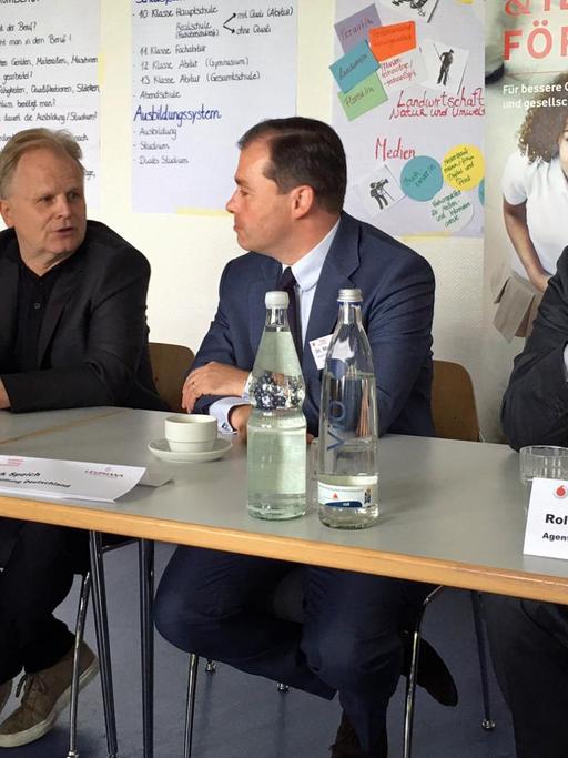 Drei Männer mittleren Alters sitzen an einem Tisch mit Mikrofonen vor sich. Sie werden gleich eine Pressekonferenz geben.