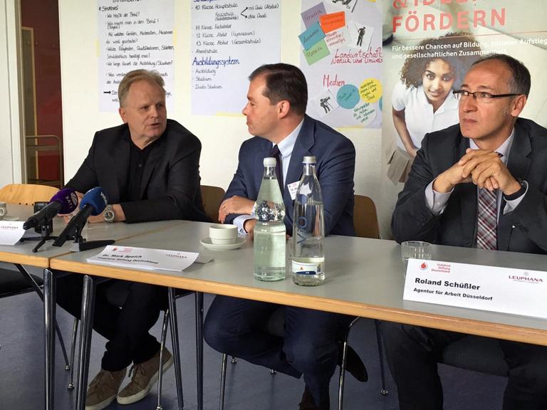 Drei Männer mittleren Alters sitzen an einem Tisch mit Mikrofonen vor sich. Sie werden gleich eine Pressekonferenz geben.