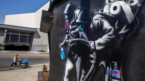 Eine Frau läuft an einem Wandgemälde von 'Hijackhart' vorbei. Es zeigt Soldaten, die desinfizieren. Los Angeles, Kalifornien, 2020.