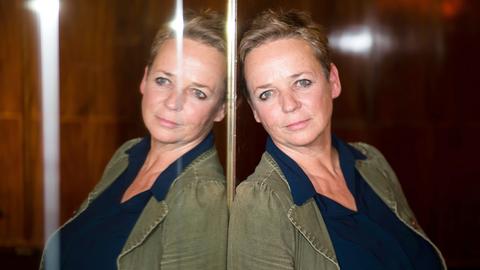 Eine mittelalte Frau mit blonder Kurzhaarfrisur lehnt sich mit dem Rücken an einen Spiegel.