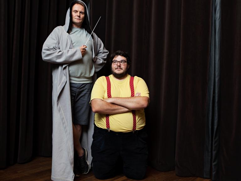 Das Bild zeigt Devid Striesow, der einen Kaputzenmantel trägt und auf einem Podest steht, einen Taktstock hält. Rechts neben ihm steht Axel Ranisch mit verschränkten Armen.