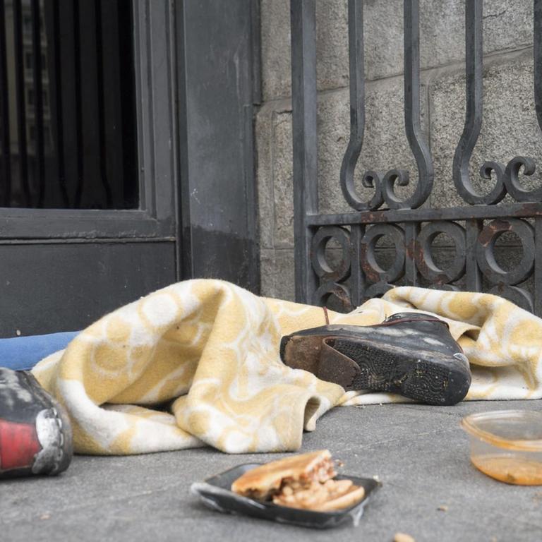 Lagerplatz eines Obdachlosen mit zerrissenen Schuhen, Essensresten und einer Decke vor einem Geschäft in Madrid.