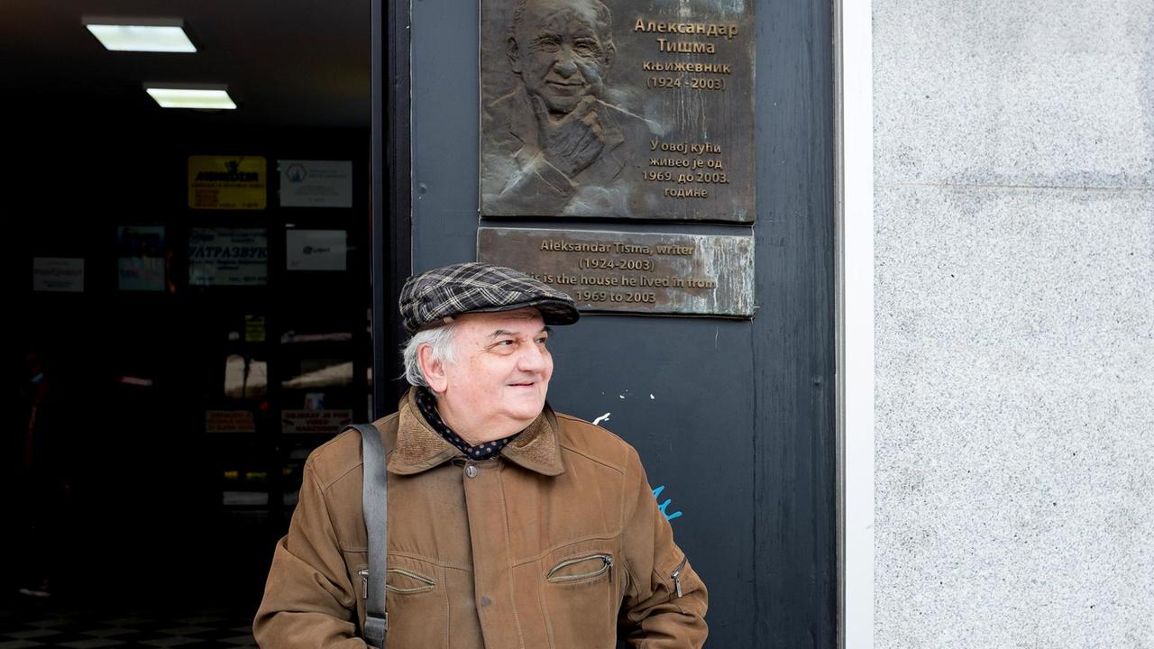 Andrej Tišma mit Schiebermütze steht lächelnd an einem Hauseingang, über ihm eine Hinweistafel zu seinen Vater