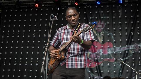 Ein Mann mit in einem kariertem Hemd steht auf einer Bühne und spielt Gitarre.