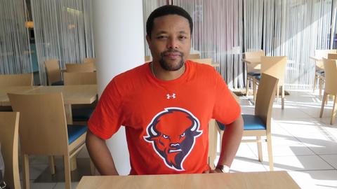 Ein Mann im orangen T-Shirt sitzt in einem leeren Restaurant und schaut lächelnd in die Kamera.