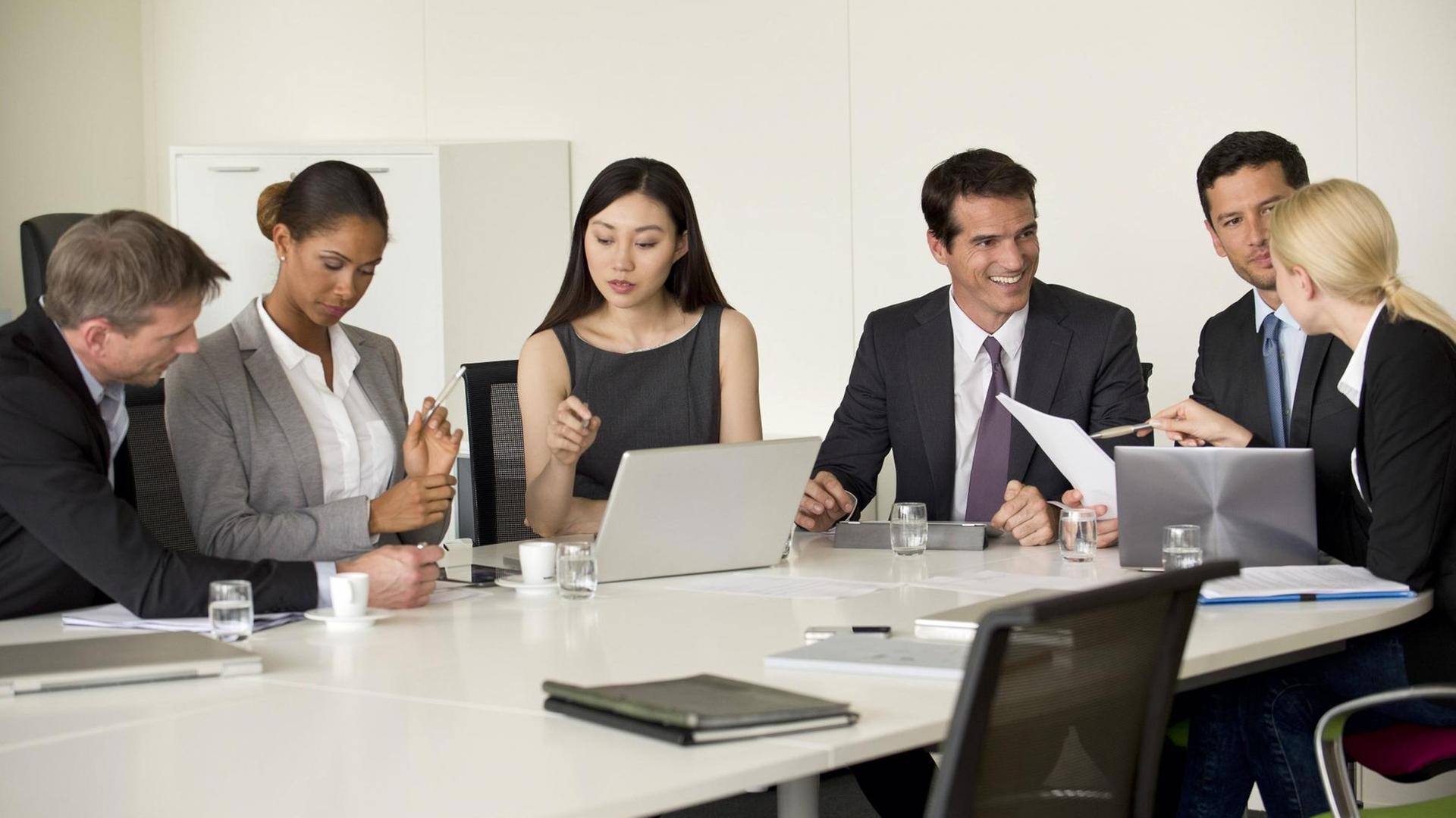Drei Männer und drei Frauen in Business-Kleidung sitzen um einen Konferenz-Tisch, auf dem Laptops stehen