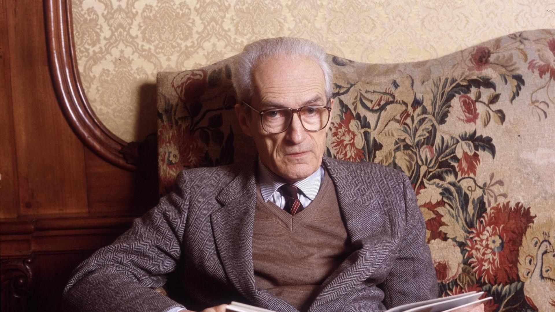 Ein alter Mann mit weißen Haaren und mit Schlips und großer Brille sitzt auf einem altmodisch wirkenden Sofa und hält ein geöffnetes Buch im Schoß.