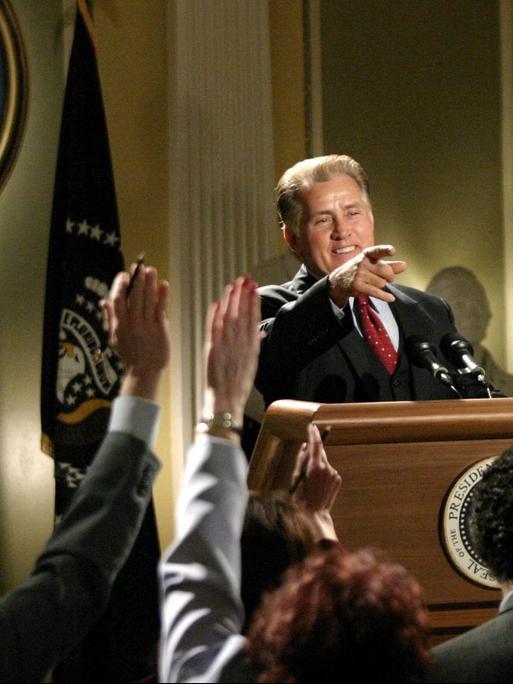 Martin Sheen steht als Präsident Josiah Bartlet in 'The West Wing' vor einem Rednerpult und zeigt auf jemanden in der Menge.