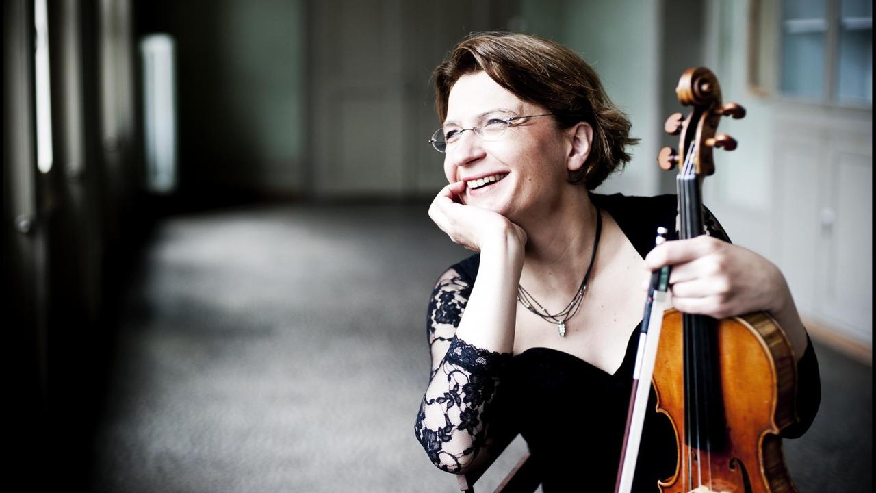 Antje Weithaas sitzt in Konzertkleidung in einem hellen Raum mit einer Fensterfront. Sie stützt ihre Geige auf die Knie.