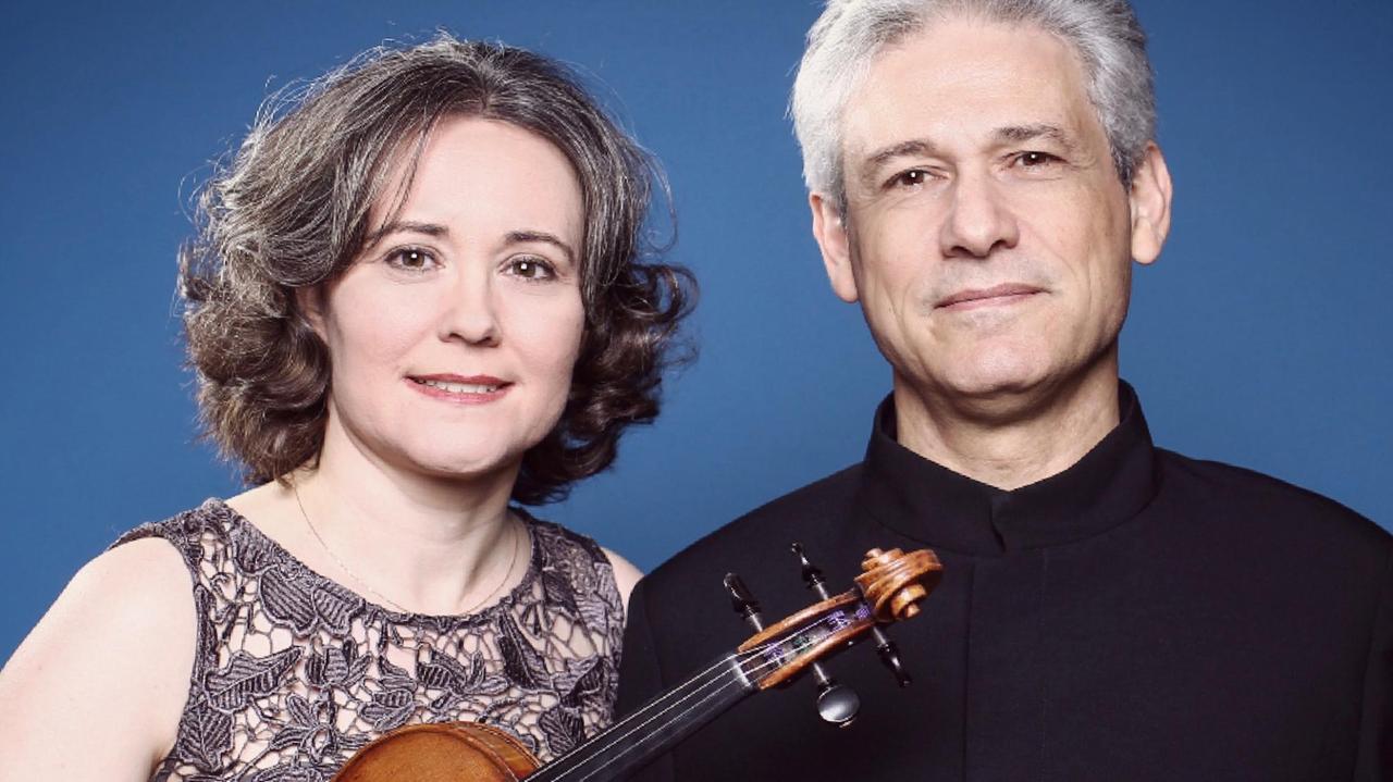 Judith Ingolfsson und Vladimir Stoupel stehen vor einem blauen Hintergrund und schauen in die Kamera. Judith Ingolfsson hält dabei ihre Geige in ihren Händen.