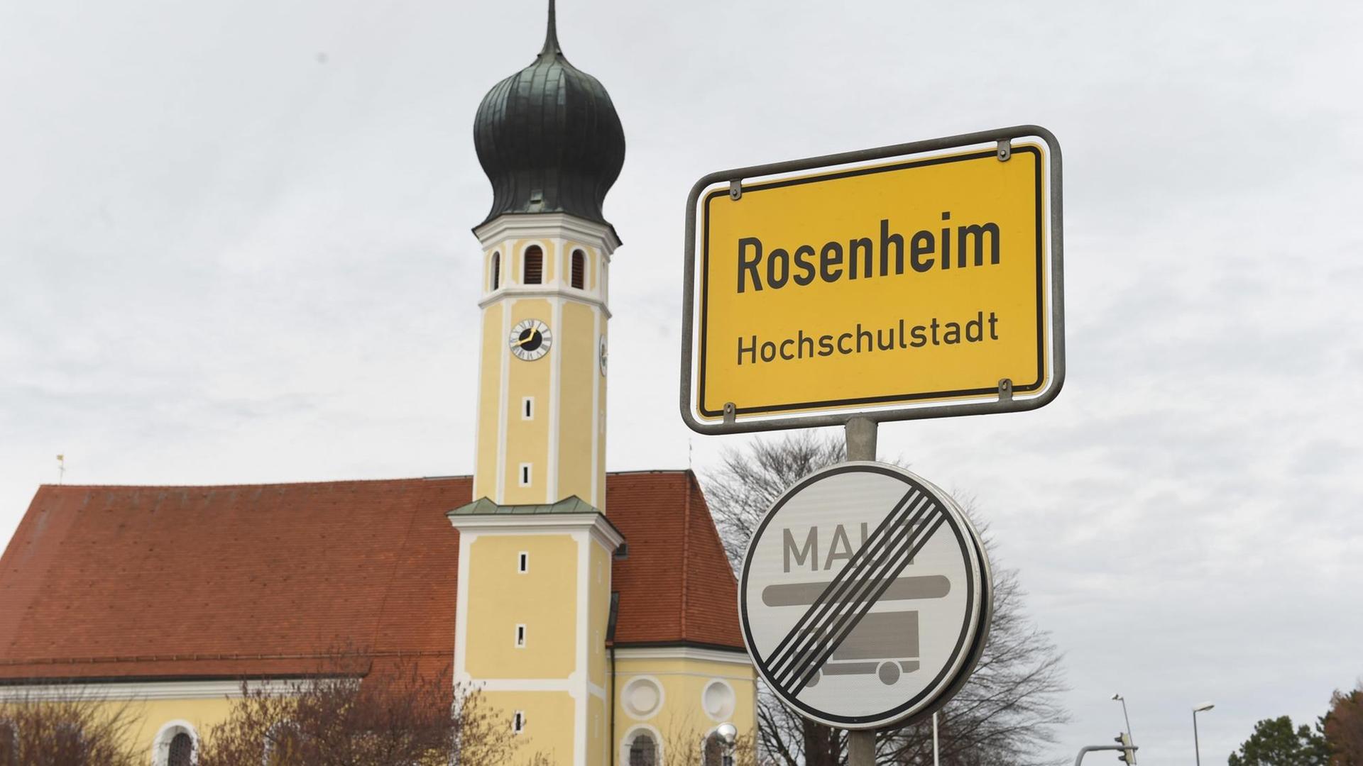 Das Ortsschild von Rosenheim mit der Kirche Heilig Blut im Hintergrund. Der Himmel ist bewölkt.