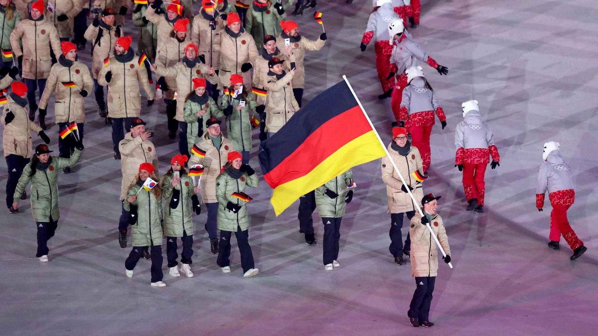 Blick von der Tribüne auf Frenzel mit der Fahne und einen Teil der deutschen Mannschaft. Daneben eine Reihe tanzender Frauen.