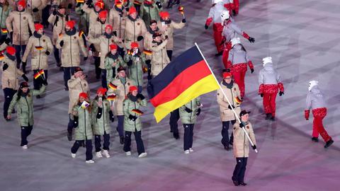 Blick von der Tribüne auf Frenzel mit der Fahne und einen Teil der deutschen Mannschaft. Daneben eine Reihe tanzender Frauen.