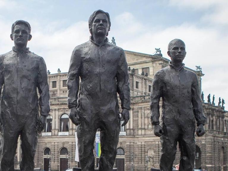 Bronzeskulpturen sind aufgebaut und stehen auf drei Stühlen (Snowden, Assange, Manning). Ganz links steht der Künstler Davide Domino.