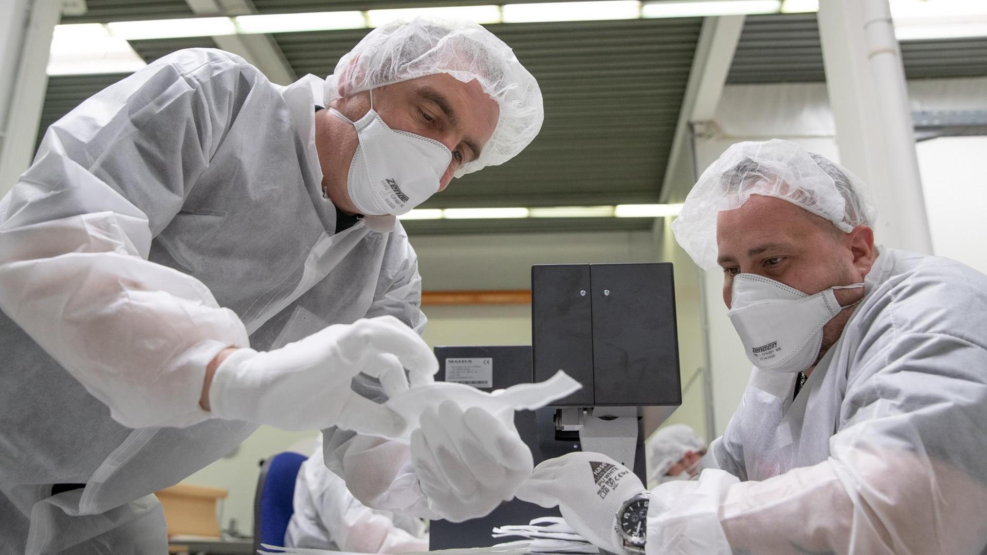Zwei Männer in weißer Schutzkleidung hantieren in einem Werkraum mit Schutzmasken.