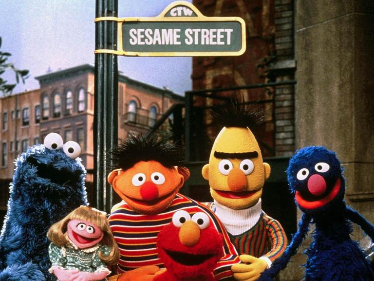Die Puppen der Kindersendung Sesamstraße, das Krümelmonster, Mariechen, Ernie, Elmo, Bert und Grobi stehen vor einem Straßenschild mit der Aufschrift "Sesame Street"