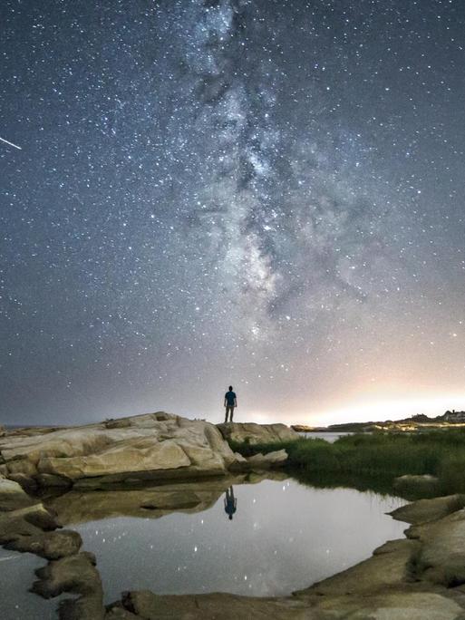 Ein Mensch steht vor einem Sternenhimmel, auf dem eine Sternschnuppe zu sehen ist.