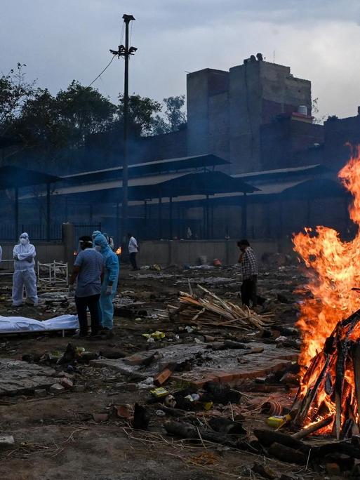 Menschen in Schutzanzügen stehen in der Nähe eines toten Angehörigen, der am Coronavirus gestorben ist, auf einem Einäscherungsgelände in Neu-Delhi. Im rechten Bildvordergrund ist ein Feuer zu sehen.
