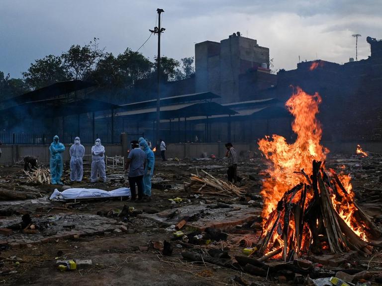 Menschen in Schutzanzügen stehen in der Nähe eines toten Angehörigen, der am Coronavirus gestorben ist, auf einem Einäscherungsgelände in Neu-Delhi. Im rechten Bildvordergrund ist ein Feuer zu sehen.