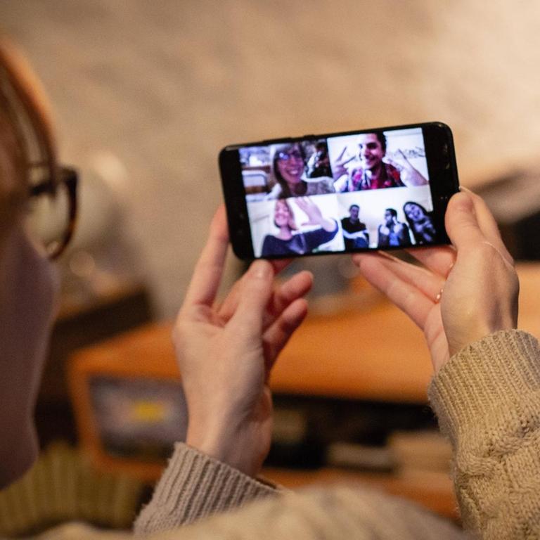 Eine Frau hat ein Smartphone der Hand, auf dem Bildschirm sind sechs Verwandte oder Freunde zu sehen, mit denen sie per Video kommuniziert.