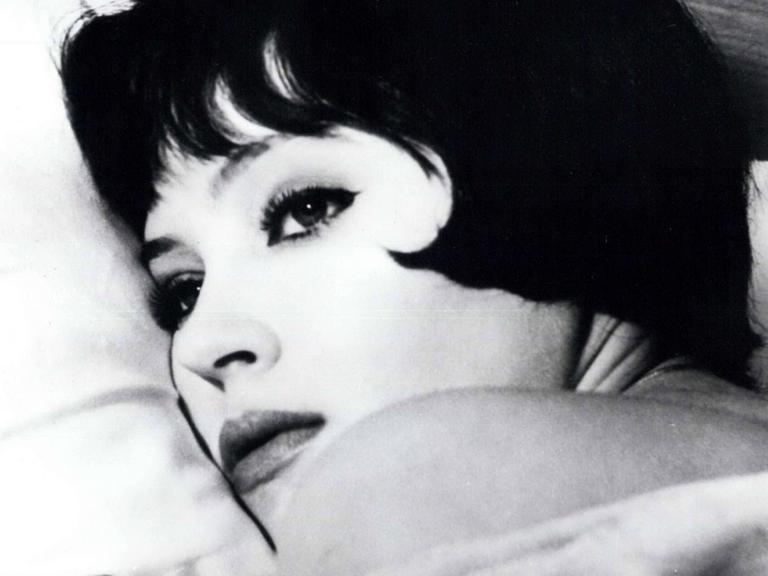 Eine Filmszene aus "Die Geschichte der Nana S." von Jean Luc Godard zeigt Anna Karina in einer Nahaufnahme auf einem weißen Kissen liegend.