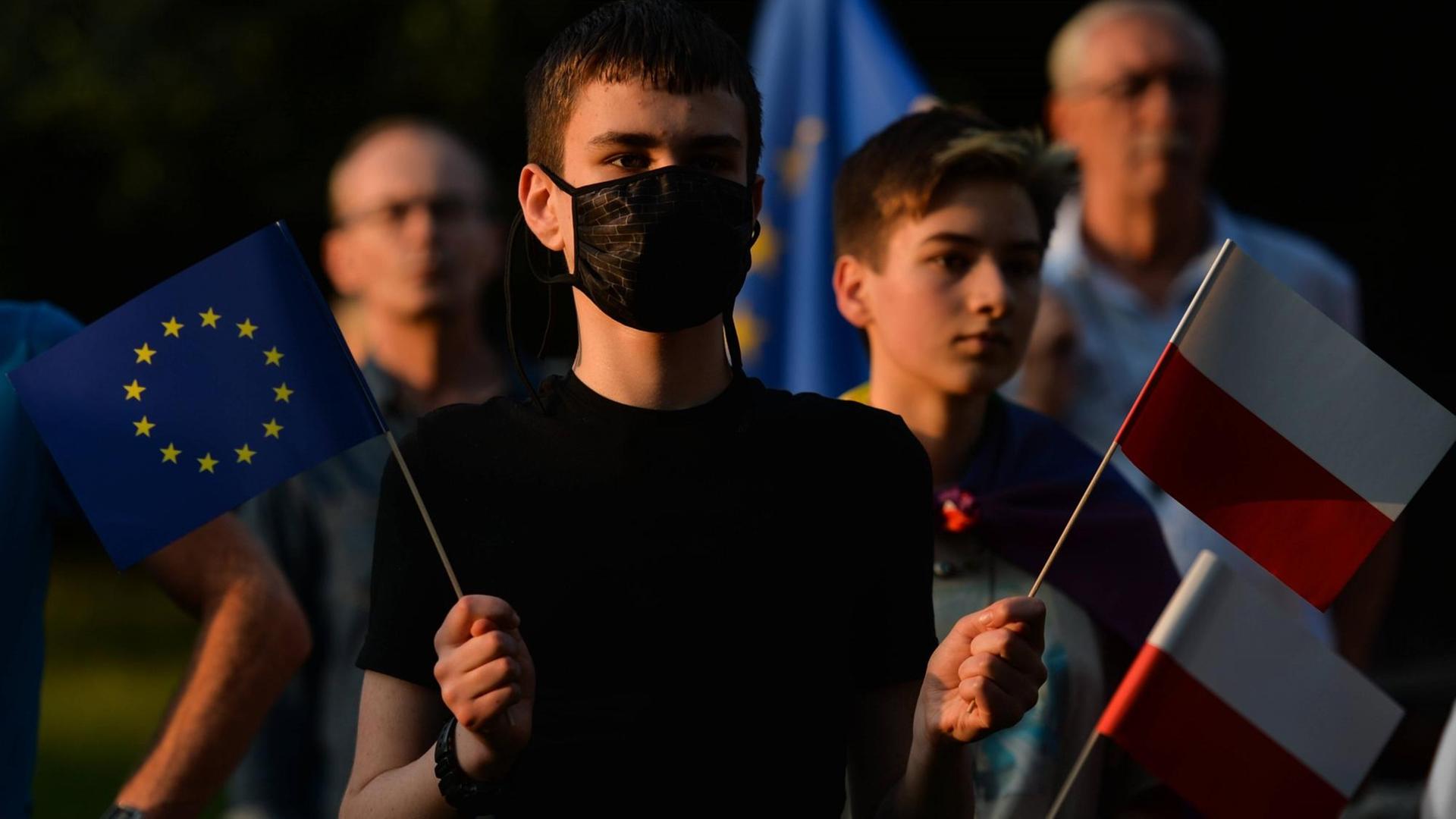 Ein junger Mann mit Mund-Nasenschtz hält bei einer Demo in Krakau eine polnische und eine Flagge der EU.