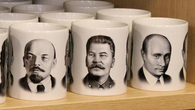 Die Gesichter Lenins, Stalins und Putins auf nebeneinanderstehenden Tassen 