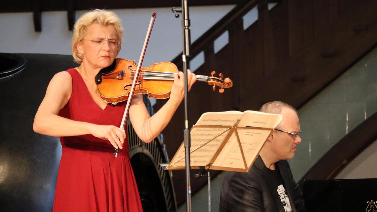 Eine Geigerin im roten Kleid spielt mit geschlossenen Augen ihr Instrument. Im Hintergrund ist ein Pianist zu sehen, der sie begleitet.
