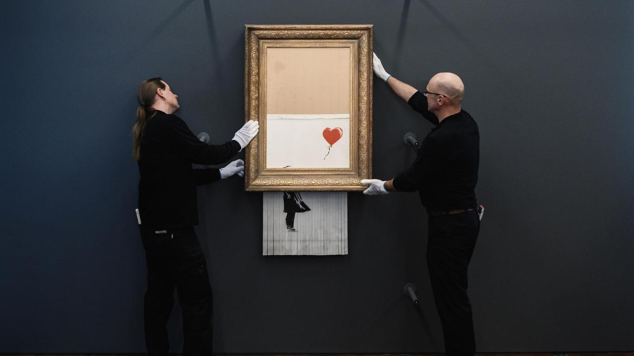 Zwei in schwarz gekleidete Museumsmitarbeiter hängen mit weißen Handschuhen Banksys Bild "Love in the Bin" an die Wand. Die Hälfte des Bildes hängt halb geschreddert aus dem goldenen Rahmen heraus.