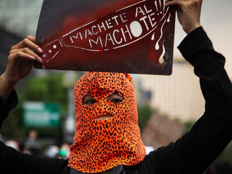 Eine Demonstrantin, in einer Leopardenmuster-Maske, bei Protesten gegen Femizide in Mexiko Stadt, mit einem Schild mit der Aufschrift "Machete all Machote!!!"