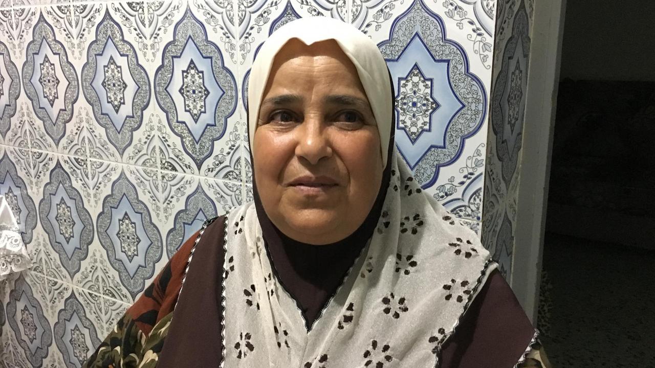 Die Tunesierin Latifa Matmati in einem großen, gemusterten Schal ist vor einer Wand mit blau-weißen Kacheln zu sehen.