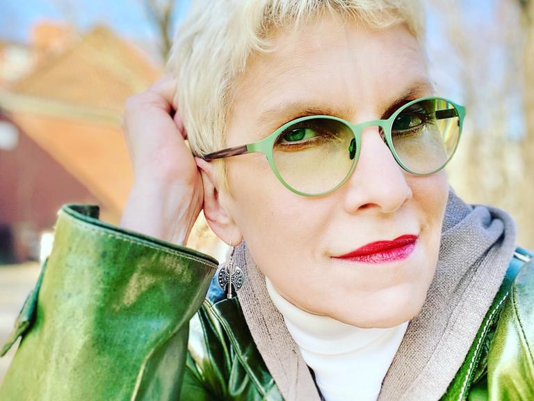 Portrait der Schriftsteller*in Else Buschheuer in grünem Mantel und mit grüner Sonnenbrille.
