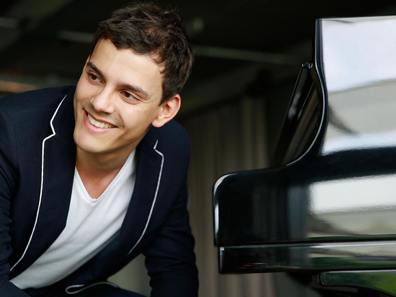Der Pianist sitzt und schaut rechts seitlich in die Kamera, er trägt ein weißes t-shirt und ein blaues Jacket. Von ihm links aus steht der schwarze glänzende Flügel, zugeklappt.