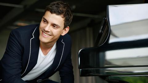 Der Pianist sitzt und schaut rechts seitlich in die Kamera, er trägt ein weißes t-shirt und ein blaues Jacket. Von ihm links aus steht der schwarze glänzende Flügel, zugeklappt.
