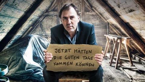 Stefan Waghubinger sitzt auf einem verstaubten Dachboden und hält ein Pappschild in den Händen, auf dem der Titel seines beuen Programms steht: "Jetzt hätten die schönen Tage kommen können".