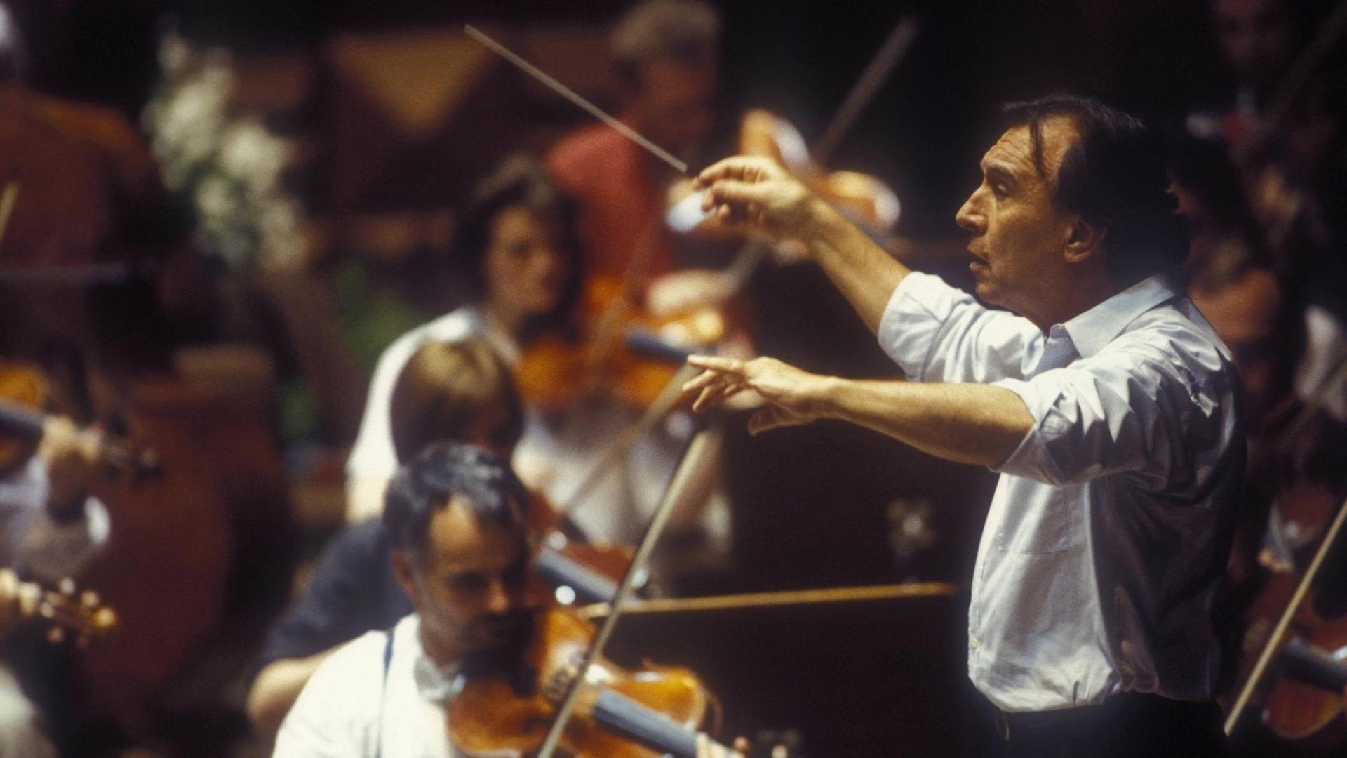 Ein Mann mit schwarzen Haaren steht mit hochgekrempelten Ärmeln seines weißen Hemdes am Dirigentenpult vor einem Orchester und hält den Taktstock in die Höhe. Ein Mann mit schwarzen Haaren steht mit hochgekrempelten Ärmeln seines weißen Hemdes am Dirigentenpult vor einem Orchester und hält den Taktstock in die Höhe.