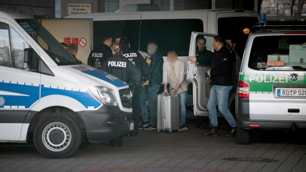 Mit Handschellen gefesselt schiebt ein Mann am Frankfurter Flughafen seinen Rollkoffer zwischen Polizisten hindurch, ein anderer trägt einen Pappkarton.
