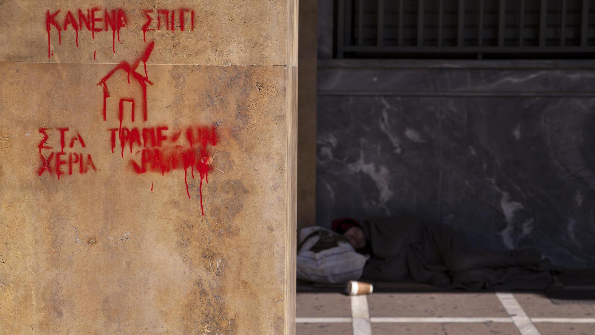 Der Schriftzug "Kein Haus in den Händen eines Bankers" steht in roter Farbe an einer Hauswand, im Hintergrund ist ein Obdachloser zu sehen