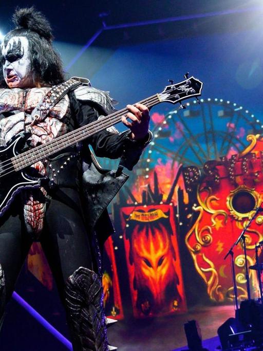 Gene Simmons, der dämonisch geschminkte und verkleidete Bassist der Band Kiss, steht in theatralischer Pose auf einer Bühne, die wie ein Horror-Zirkus aussieht.