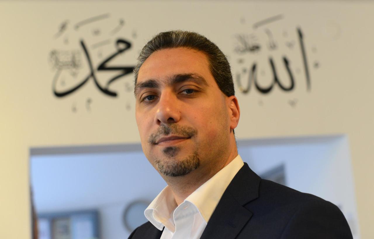 Sadiqu Al-Mousllie im schwarzen Sakko und weißem Hemd vor einem Türeingang mit arabischen Schriftzügen. Er blickt in die Kamera.