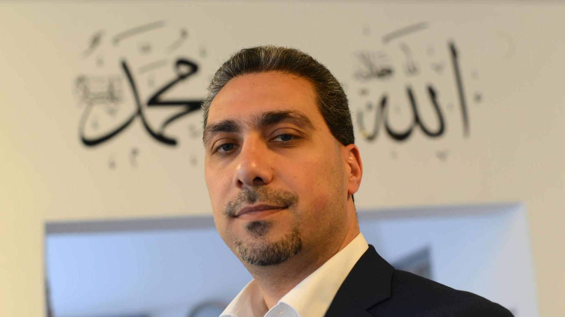 Sadiqu Al-Mousllie im schwarzen Sakko und weißem Hemd vor einem Türeingang mit arabischen Schriftzügen. Er blickt in die Kamera.