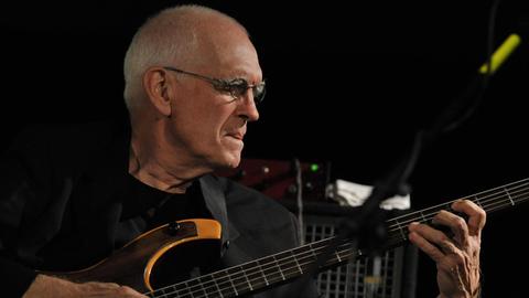 Ein älterer Mann mit Brille und kurzem, weißem Haar spielt auf einem E-Bass und schaut konzentriert auf das Griffbrett