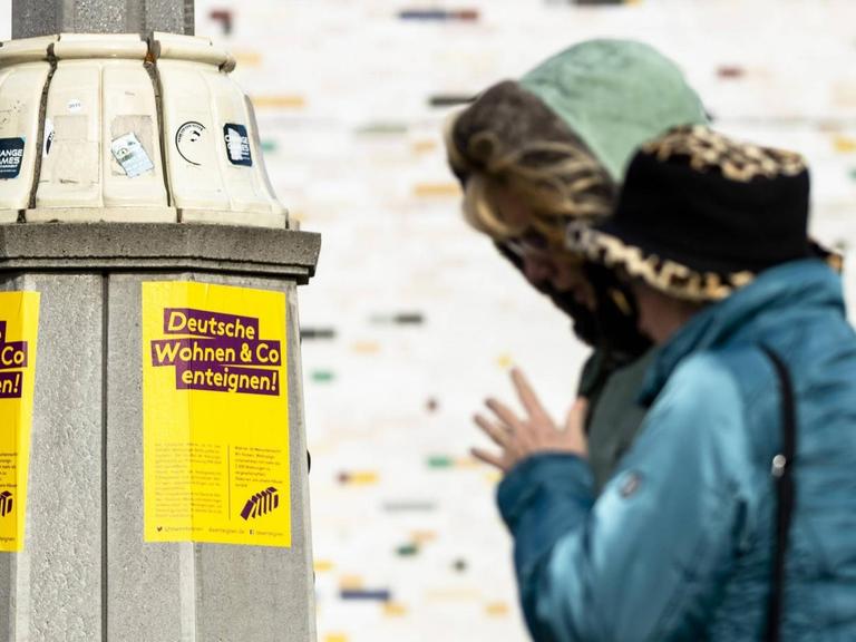 An einem Laternenmast hängt ein Aufkleber mit dem Schriftzug "Deutsche Wohnen Enteignen", im Hintergrund läuft ein älteres Ehepaar.