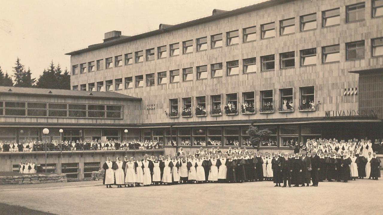 Viele Frauen und Männer stehen auf dem Vorplatz eines großen, mehrteiligen Gebäudes
