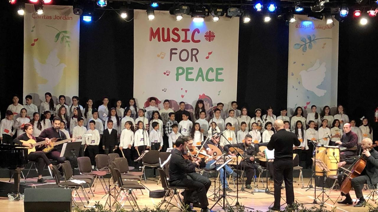Auf einer Bühne steht ein Chor mit vielen Kindern. Im Vordergrund ist ein Orchester. Im Hintergrund ist ein Transparent mit der Aufschrift "Music for Peace" zu sehen.